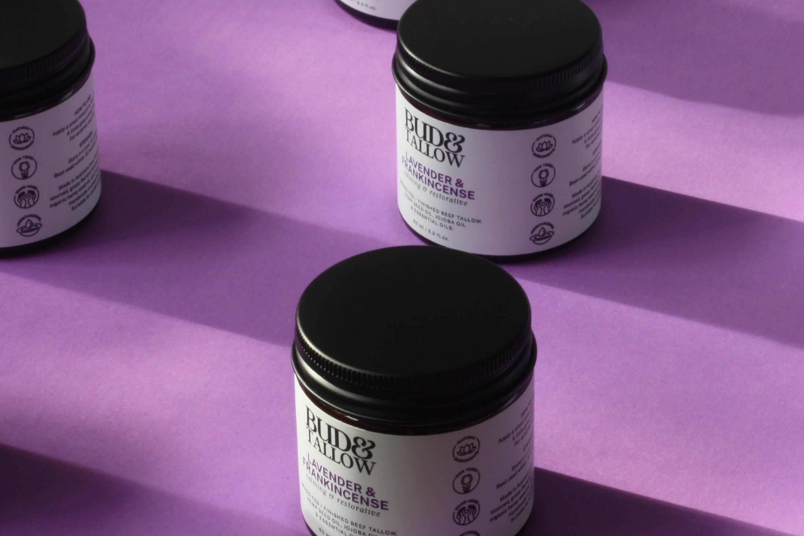 Jars of a grass fed tallow balm lavender & frankincense with black lids on a purple surface